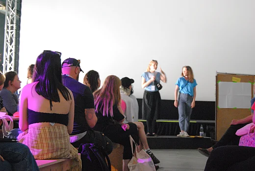 in der linken Bildh&auml;lfte Menschen in einem Raum von hinten, rechts im Hintergrund zwei Menschen auf einer B&uuml;hne