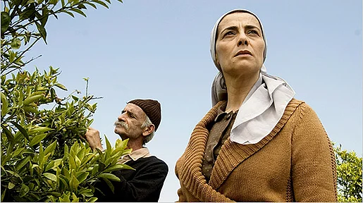 Szenenbild: Im Hintergrund steht ein alter Mann an einem Zitronenbraum. Im Vordergrund steht eine der Protagonistinnen und schaut in die Kamera