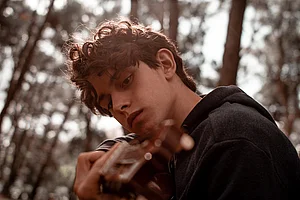 Ein Junge spielt Gitarre, hinter ihm sieht man Wald. 