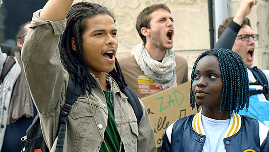 Zwei Teenager sind im Vordergrund zu sehen, wie sie die F&auml;uste heben und Parolen rufen. Im Hintergrund stehen weitere rufende Protestierende, auf einem Plakat ist das Wort ZAD zu lesen.
