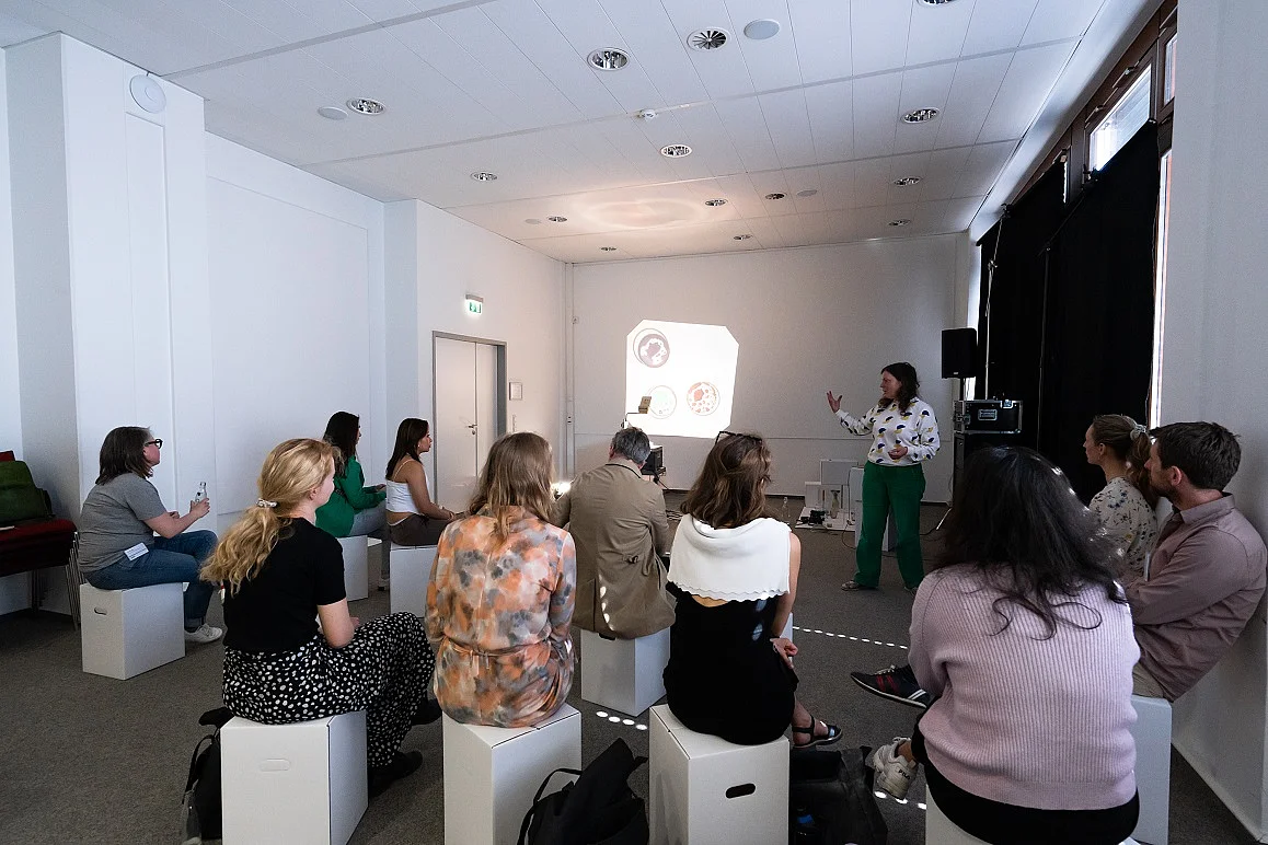 Menschen auf Pappschemeln sitzend in einem gro&szlig;en Raum, von hinten aufgenommen, sie blicken auf eine Leinwand
