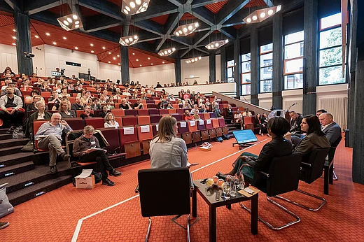 Ein gro&szlig;er, h&ouml;rsaal&auml;hnlicher Raum mit vielen besetzten Stuhlreihen. Im Vordergrund, von hinten aufgenommen, Menschen auf St&uuml;hlen in einer Gespr&auml;chsrunde