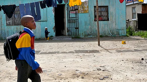 Szenenbild aus Südafrika: Ein kleiner Junge geht an einer türkisfarbenen Wellblechhütte vorbei, vor der Wäsche auf der Leine hängt