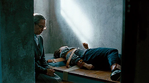 Szenenbild: eine junge Frau liegt angeschnallt auf einer Holzpritsche, eine alte Frau sitzt daneben und liest