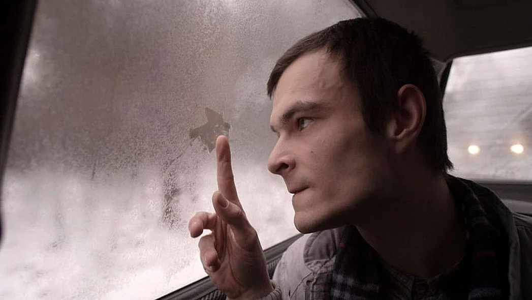 Ein junger Mann sitzt im Auto und schaut aus dem Fenster. Mit seinem Finger wischt er an der beschlagenen Scheibe.