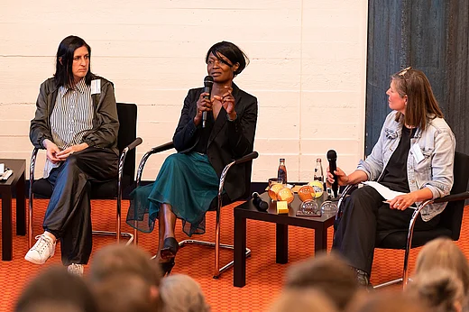 Drei Menschen auf einer B&uuml;hne, im Vordergrund unscharf K&ouml;pfe von Zuh&ouml;rer*innen, Sheri Hagen spricht in ein Mikrofon