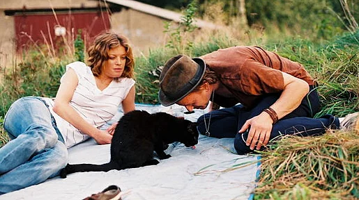 Szenenbild: Ein Mann und eine Frau sitzen auf einer Decke um eine schwarze Katze herum
