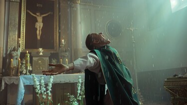 Ein junger Mann in Kleidung eines Priesters steht mit ausgebreiteten Armen in einer Kirche und sieht mit euphorischem Gesichtsausdruck nach oben.