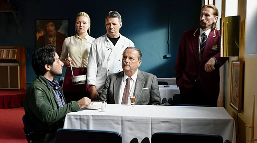 Szenenbild: Szene im Restaurant: Wikström und Khaled an einem Tisch sitzend, dahinter steht das restliche Personal