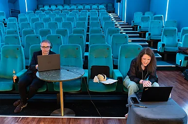 Leopold Gr&uuml;n und Lisa Hau&szlig;mann mit Computern in einem Kinosaal mit t&uuml;rkisfarbenen Sesseln