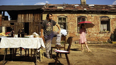 Szenenbild: Ein gedeckter Tisch im Hof eines etwas baufälligen Bauernhofs, ein Mann steht am Tisch, im Hintergrund ein Mädchen mit Schirm
